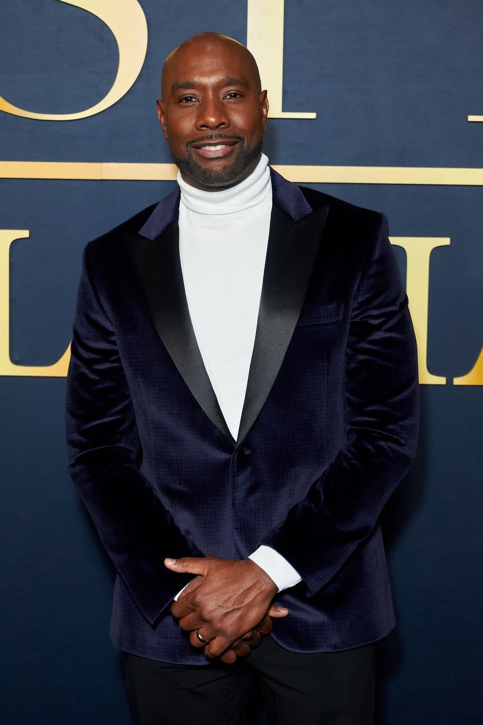 morris chestnut posing for a picture with a smile on his face, wearing a white roll neck jumper and navy blue suit jacket