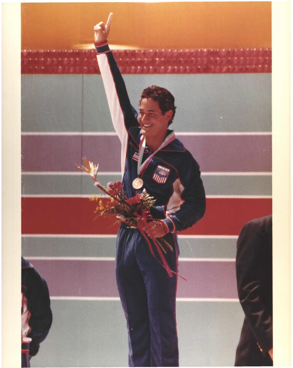 Holding a bouquet, Greg Louganis raises his arm and points in the air, looking exultant.