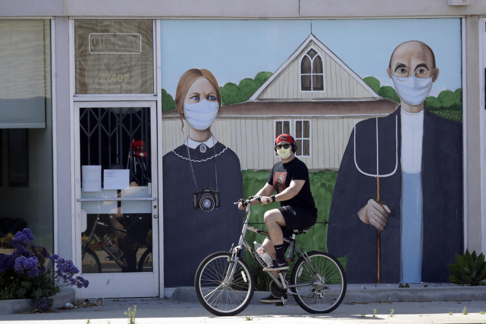 FILE - In this April 30, 2020, file photo, a person wearing a mask cycles past a recently painted mural with a depiction of artist Grant Wood's famed "American Gothic" painting, but with the subjects wearing masks, in Torrance, Calif. California's major deal for hundreds of millions of N95 respirator masks hit a delay in its federal certification process, Gov. Gavin Newsom said Wednesday, May 6, 2020, as he promised details of the contract would soon be made public. (AP Photo/Marcio Jose Sanchez, File)