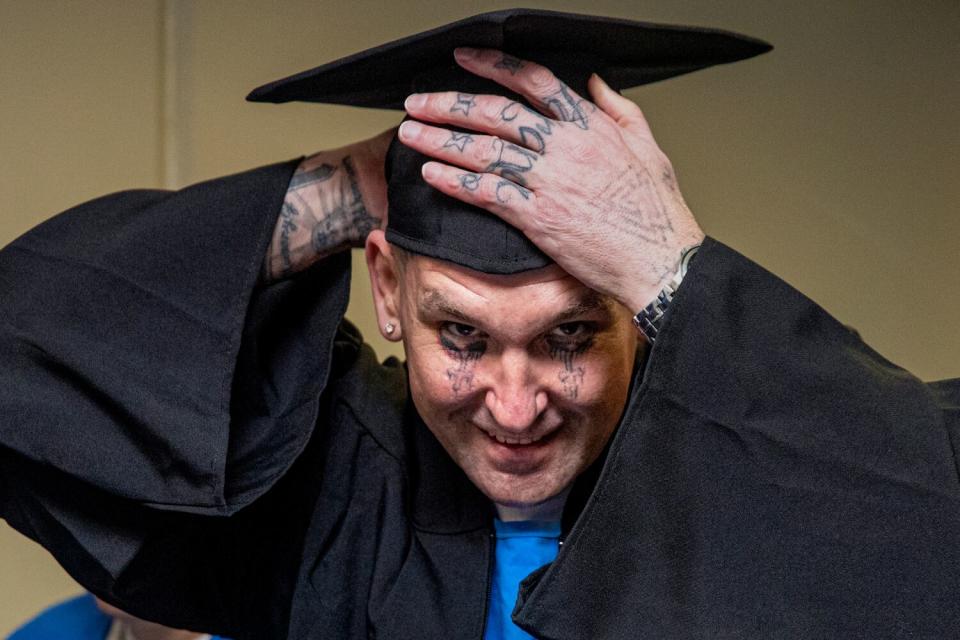 A man with facial and hand tattoos looks into the camera as he adjusts his black graduation cap.