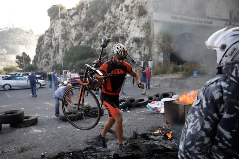 A cyclist carries his bicycle as he crosses a burning barricade on the highway, during ongoing anti-government protests at Nahr El Kalb