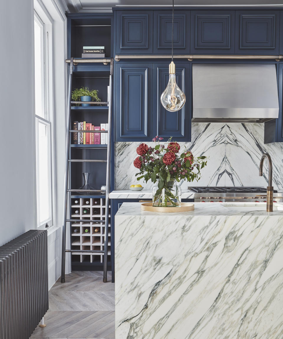 A kitchen island clad with white marble, with blue cabinetry on the rest of the kitchen, wine rack and ladder to top shelves