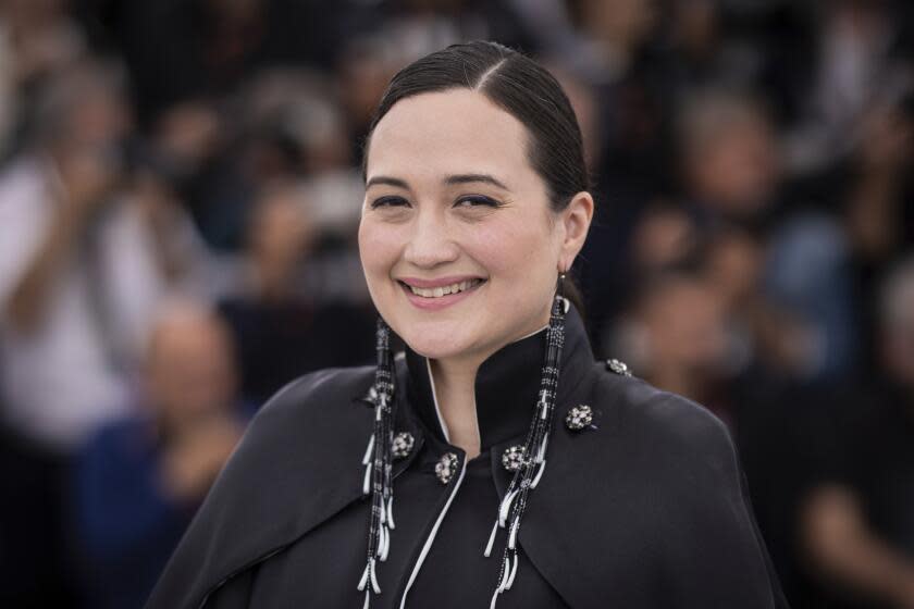 Lily Gladstone smiling in a black outfit and earrings in front of a blurred out crowd