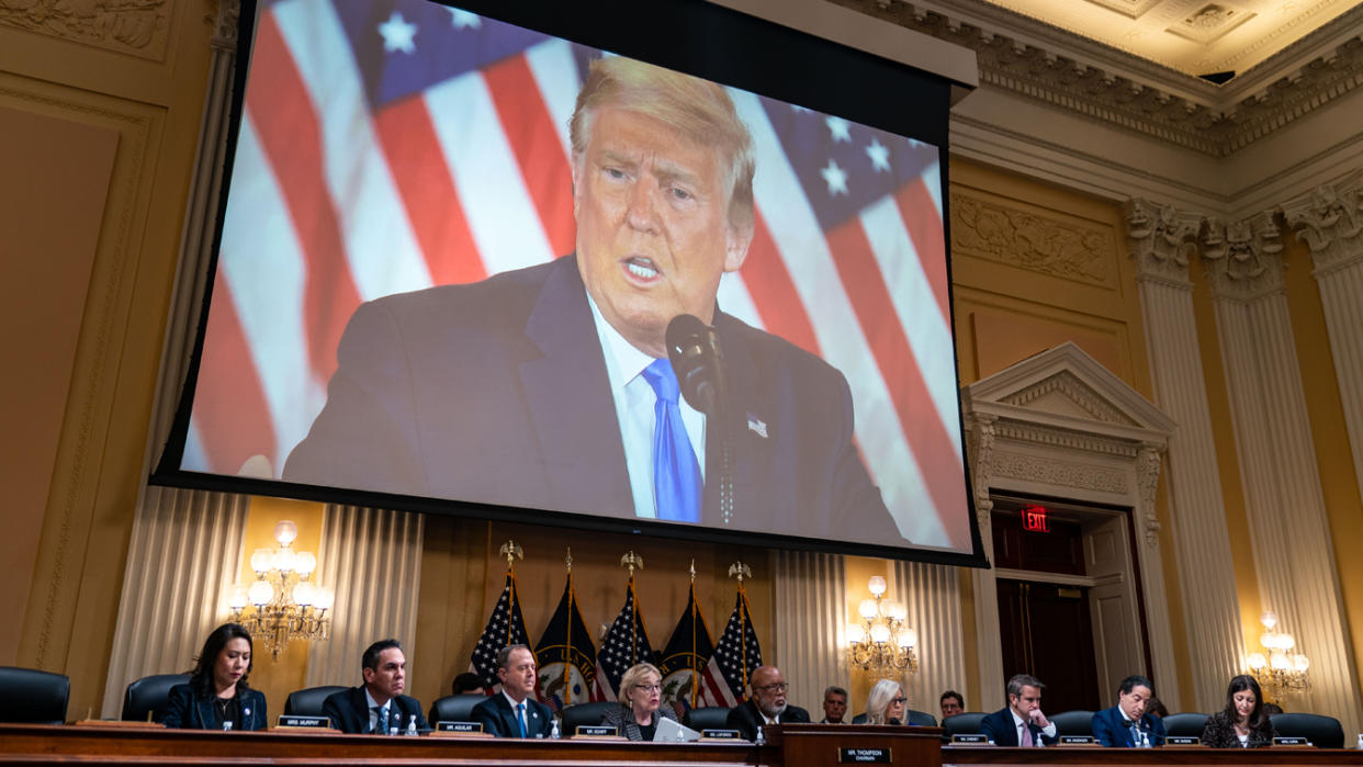 Former President Donald Trump is seen at the microphone in front of a bank of U.S. flags, with the House panel at their desks below in the Cannon House Office Building.