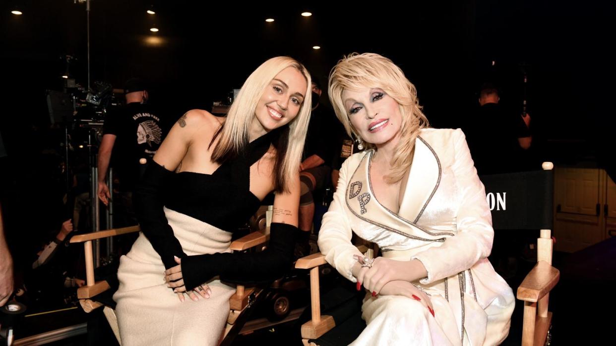 miley cyrus and dolly parton leaning in for a photo as they sit together on stage chairs