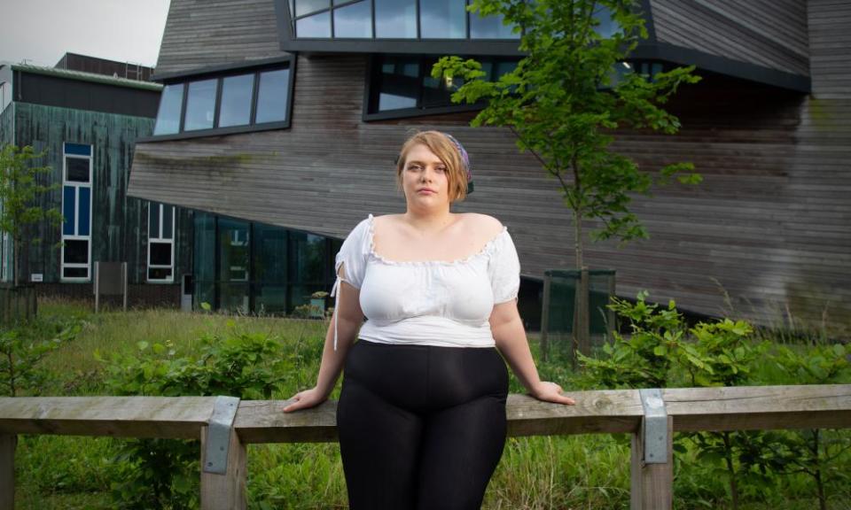 Emily Smith, con una camisa de manga corta, se apoya contra una baranda de madera frente a un edificio moderno de madera.