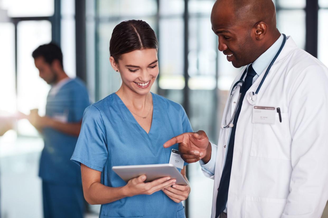 Smiling female physician assistant holding tablet while listening to cheerful male doctor with a blurred background of large windows in a hospital