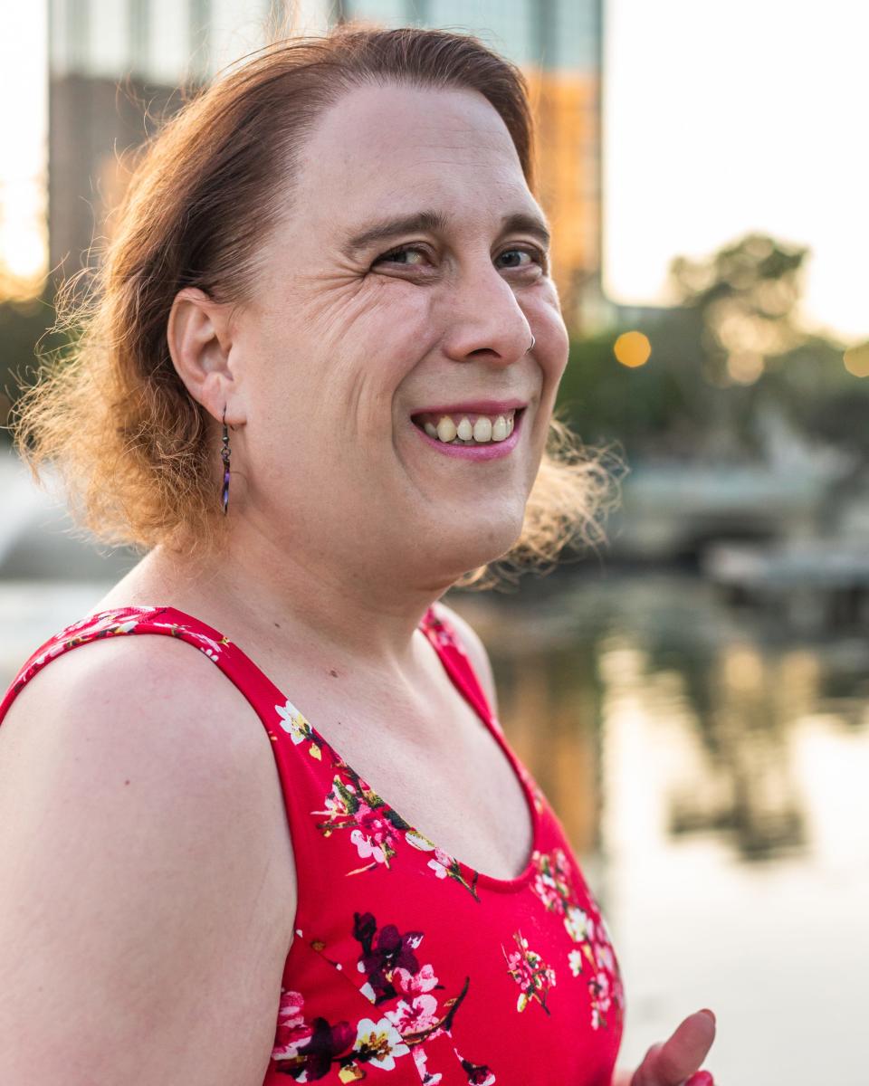 Amy Schneider smiles while wearing a red floral dress and posing outside near a cityscape.