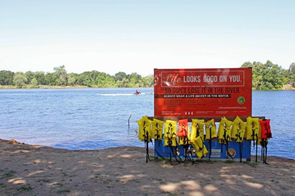 El bombero Ross Palmerton del Departamento de Bomberos de Sacramento –quien puede verse al fondo de la imagen que tiene en primer plano un estante de chalecos salvavidas y un letrero– auxilia en un simulacro de rescate en Tiscornia Beach, parte del río American, el martes 23 de mayo de 2023. Antes del fin de semana de Memorial Day, funcionarios municipales de la oficina de gestión de emergencias, el departamento de bomberos y el departamento de juventud, parques y enriquecimiento comunitario organizaron demostraciones que incluían rescate de ahogamiento y administración de RCP.