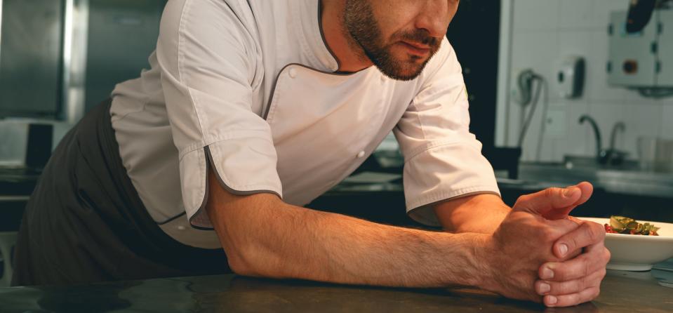 Chef leans on a kitchen counter, looking pensive while observing a dish in a commercial kitchen