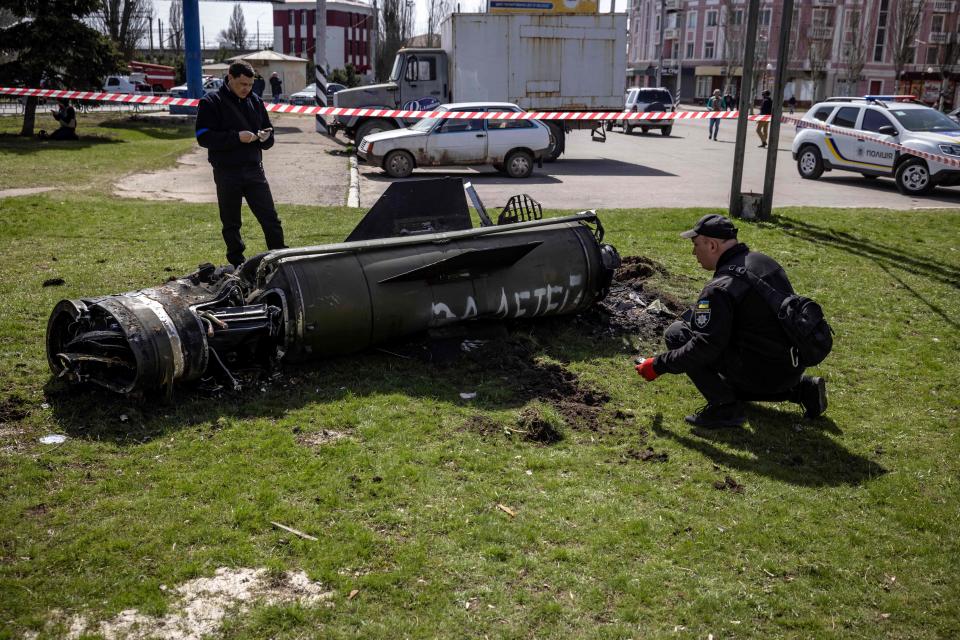 Two men in black clothes, one with a badge on this sleeve, inspect the remains of a large rocket on a grassy area near a paved area with parked vehicles and buildings in the background.