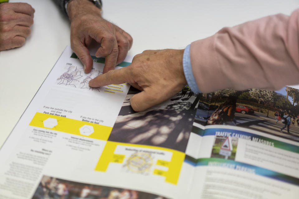 The mayor, wearing a pink jacket and blue shirt, points to a diagram in a city brochure in English in a section on what it calls Traffic Calming Measures. 