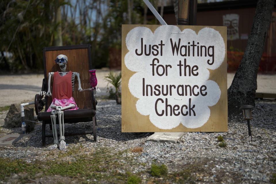 A skeleton in sunglasses sits beside a sign reading "Just waiting for the insurance check," outside the closed Kona Kai Motel on Sanibel Island, Fla., Thursday, May 11, 2023. In Sanibel, the lingering damage is not quite as widespread as in Fort Myers Beach, but many businesses remain shuttered as they are repaired and storm debris is everywhere. Seven local retail stores have moved into a shopping center in mainland Fort Myers, hoping to continue to operate while awaiting insurance payouts, construction permits, or both before returning to the island. (AP Photo/Rebecca Blackwell)