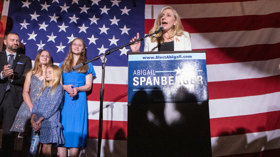 Rep. Abigail Spanberger speaks into a microphone at a podium in front of an American Flag with her husband and three daughters standing to the side..