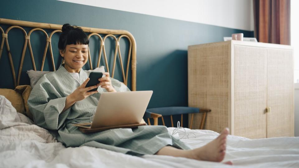 smiling woman with laptop using smart phone on bed at home