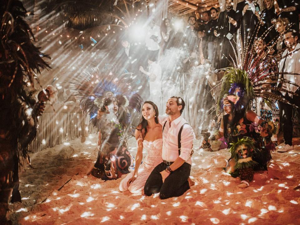 A bride and groom kneel in the sand as dancers perform for them under sparkling lights.