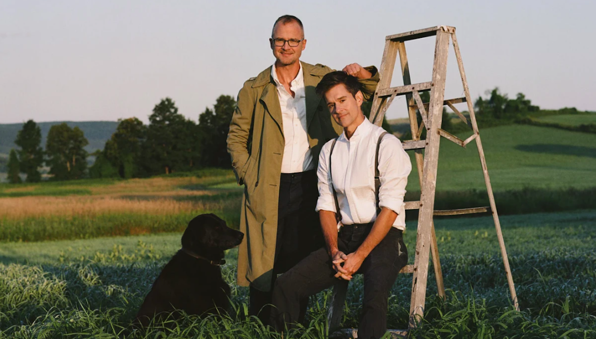 two men leaning on ladder with black dog nearby and rolling hills in the background