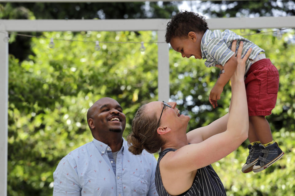 In this Thursday, May 28, 2020 photo, Kerwin Robinson looks on as his wife, Emma Robinson, lifts their son Tristan, 2, while being photographed at their home in Shoreline, Wash. Emma, a pharmacist at a Seattle hospital, and Kerwin, a software technology manager, decided to keep their son in daycare during the coronavirus outbreak. Emma Robinson figured the daycare was no more risky than her job as an essential health care worker. (AP Photo/Elaine Thompson)