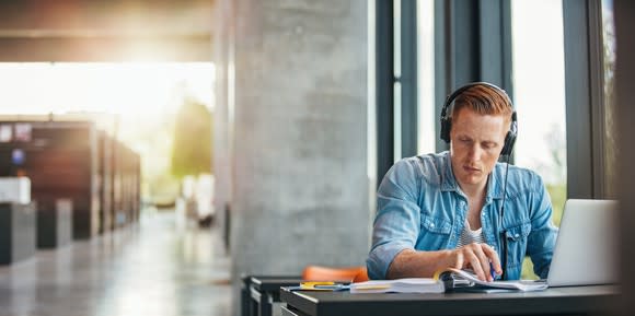 Student sitting at a table in a with a book, headphones, and a laptop.