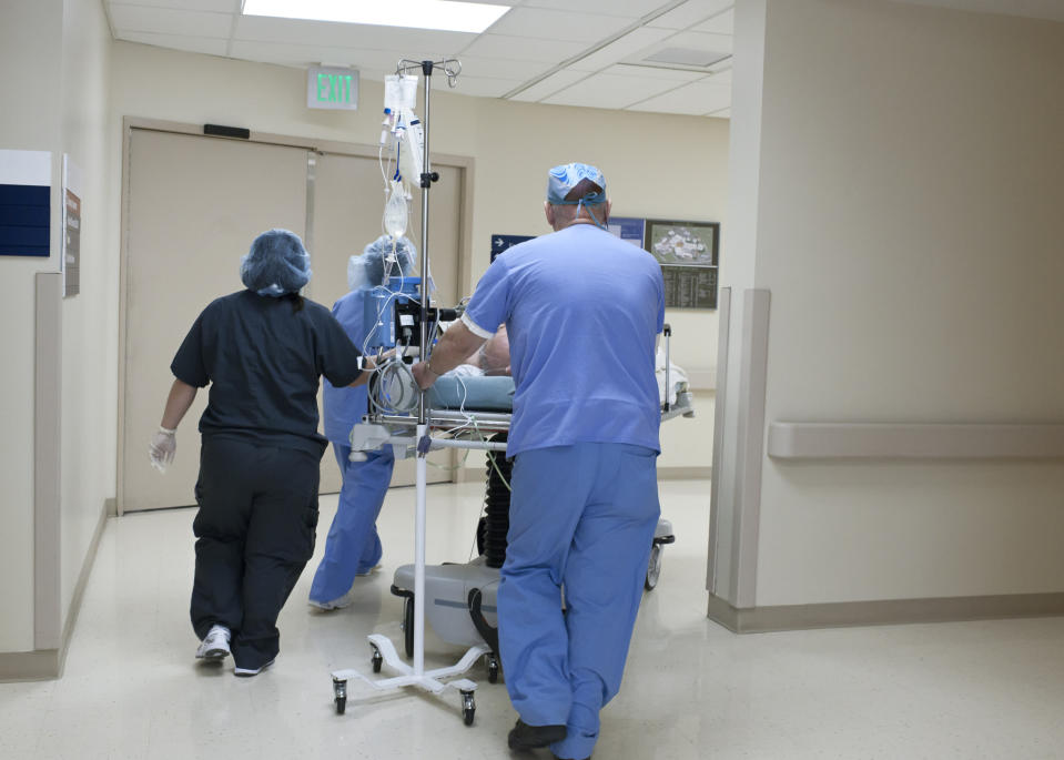 Medical team in scrubs and protective gear push a patient on a hospital bed down a hallway towards double doors