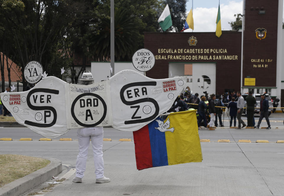 A man wearing large angel wings that read in Spanish: "S.O.S. True Peace," stands outside the General Santander police academy where a bomb exploded on campus in Bogota, Colombia, Thursday, Jan. 17, 2019. While authorities had yet to suggest who was behind the attack, and no armed group claimed responsibility, attention was focused on leftist rebels from the National Liberation Army, which has been stepping up attacks on police targets amid a standoff over how to re-start stalled peace talks. (AP Photo/John Wilson Vizcaino)