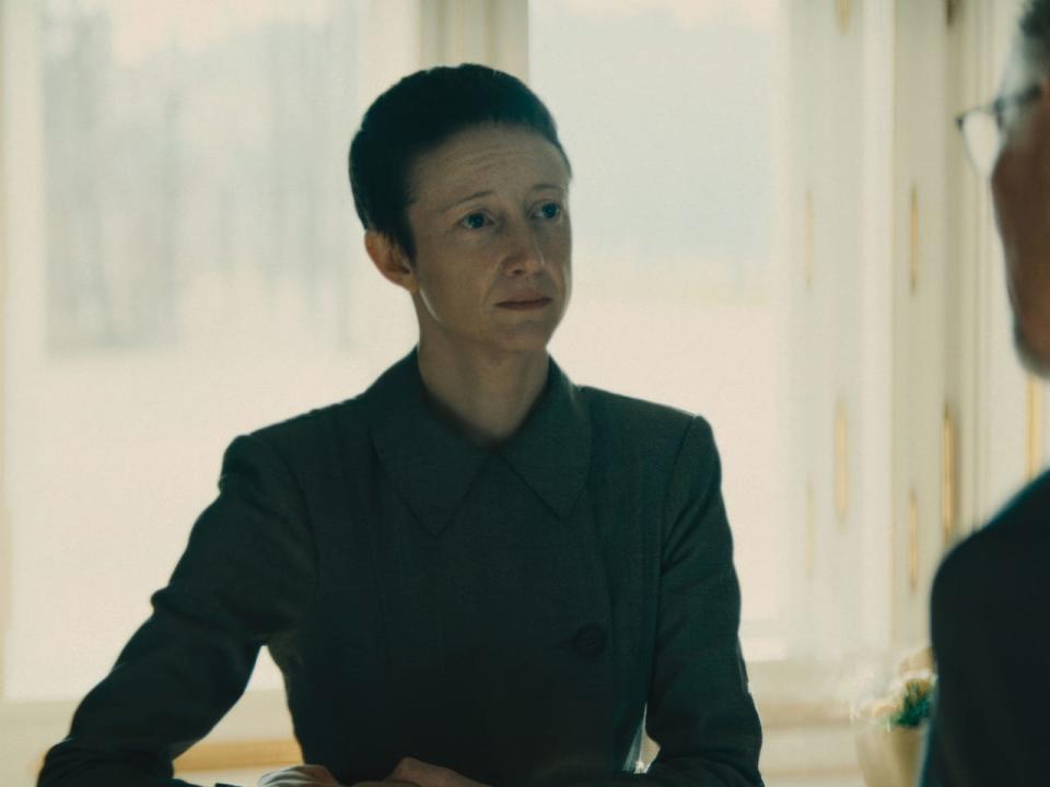 andrea riseborough in the regime, wearing a high collared professional shirt and with close cropped hair, looking at a man in the foregroudn