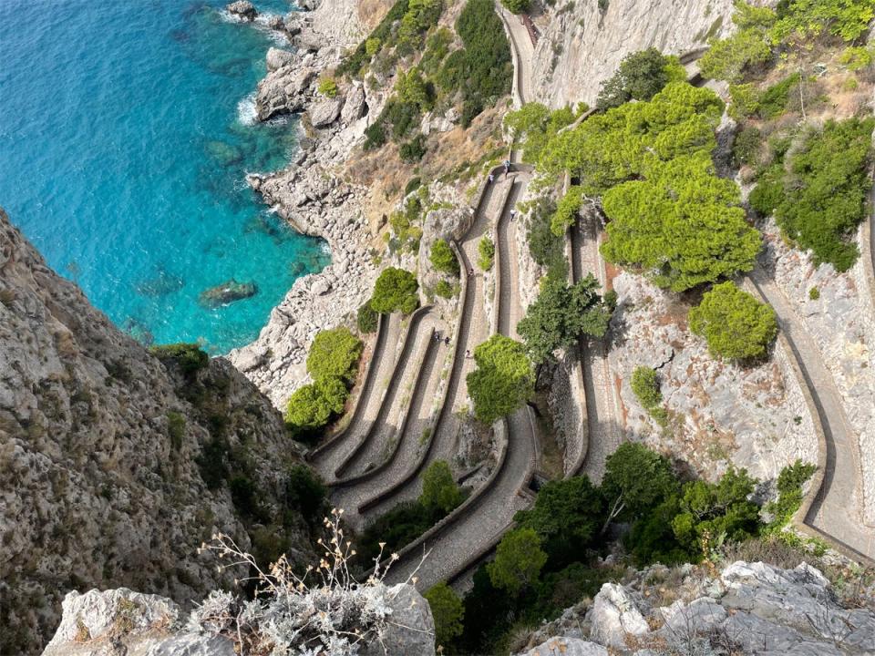 Bird's-eye-view of trails on Capri Island, surrounded by rock formations, bright-blue sea, and green trees