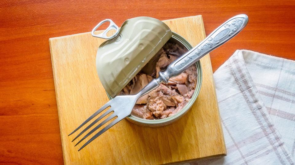 An open tuna box on a wooden board, with a fork and a towel, on a wooden table.