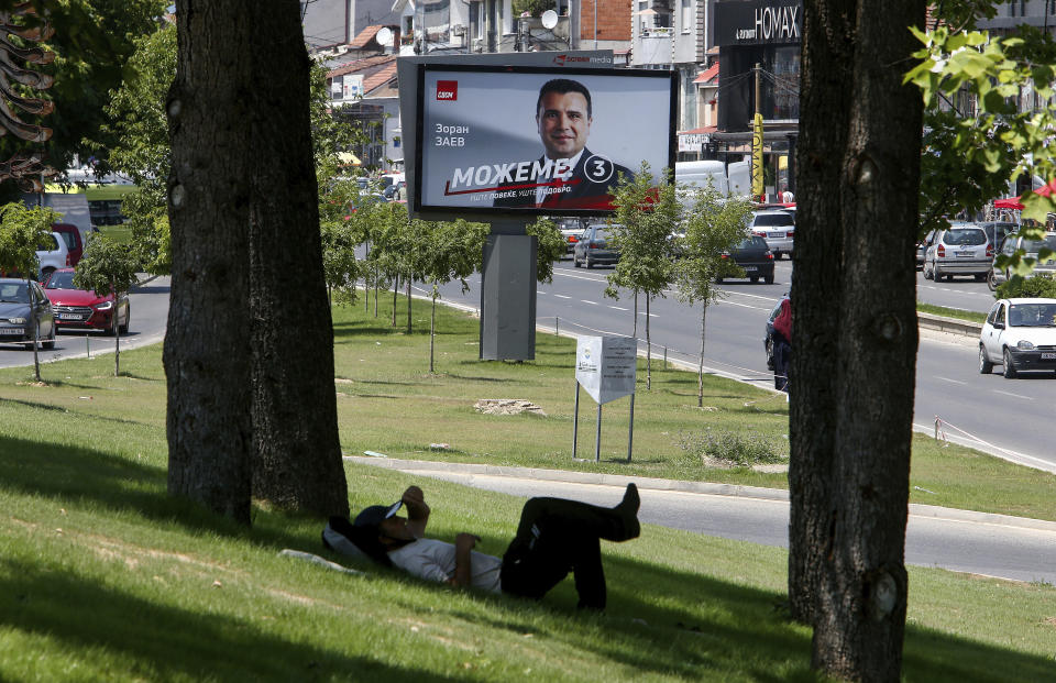 A man rests under a tree near an electoral poster of the leader of the ruling Social Democrats Zoran Zaev, in a street in Skopje, North Macedonia on Saturday, July 11, 2020. North Macedonia holds its first parliamentary election under its new country name this week, with voters heading to the polls during an alarming spike of coronavirus cases in the small Balkan nation. (AP Photo/Boris Grdanoski)