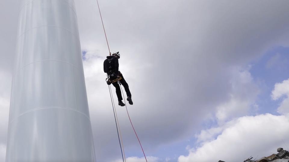 A shot looking up at Sardo hanging from a cable on the side of a wind turbine with clouds in the background.