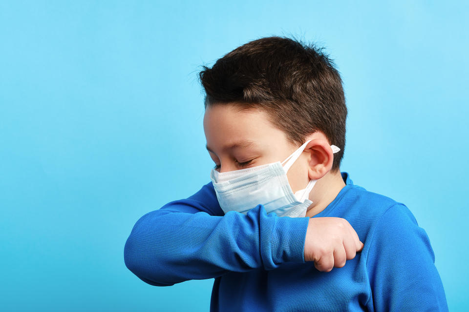 A boy wearing a medical mask coughs or sneezes into his arm. (Photo via Getty Images)