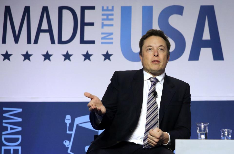 Billionaire Elon Musk, his right hand extended outward as if holding a tray, addressed a business conference while sitting in front of a backdrop that reads "Made in the USA."