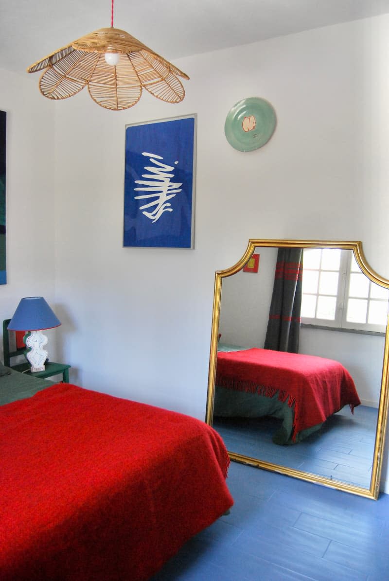 Mirror leans against white wall with blue artwork in bedroom with rattan hanging light and blue floor.