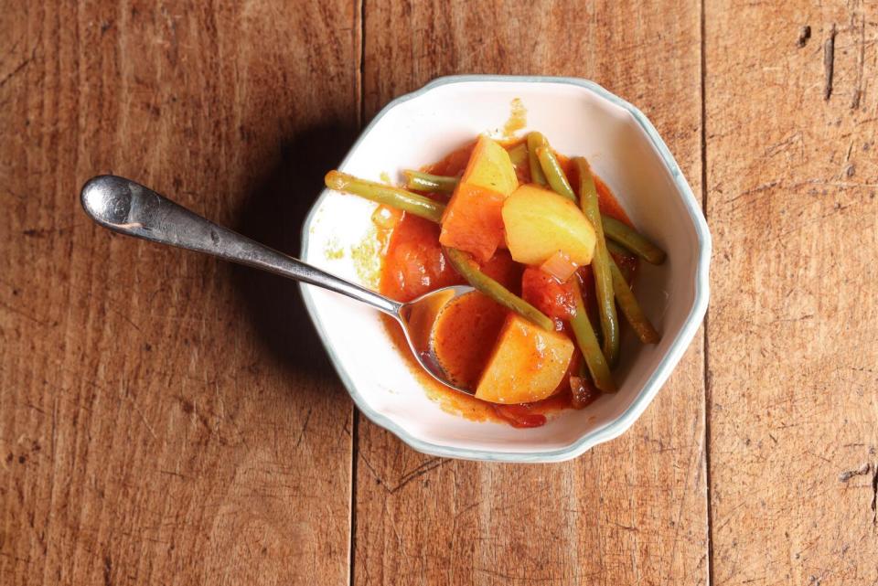 A spoon rests in a white bowl filled with fasolakia, a greek dish with green beans, potatoes, tomatoes, onions and dill.