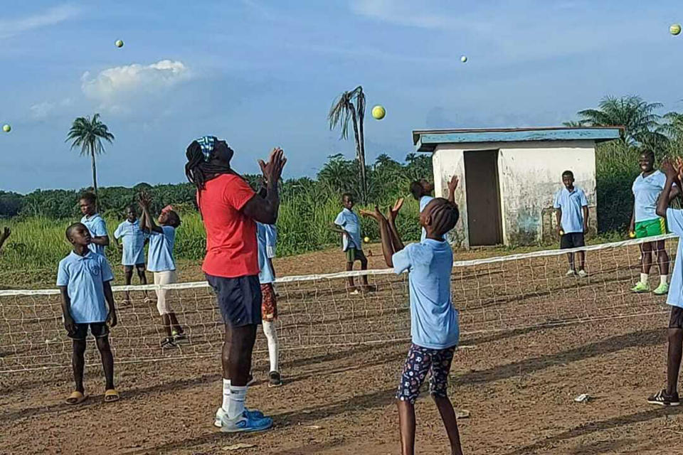 In this photo provided by Sam Jalloh, wearing a red shirt, children in Sierra Leone participate in a tennis lesson in October 2021. Jalloh zig-zagged across West Africa in 2021 coaching tennis to kids. He took with him rackets, balls and a cellphone loaded with photos and videos of a pro player in action to inspire the youngsters. The player was new U.S. tennis star Frances Tiafoe. The move to encourage young Africans by showing them images of Tiafoe, an American with Sierra Leone heritage, has paid off big time. (Sam Jalloh via AP)