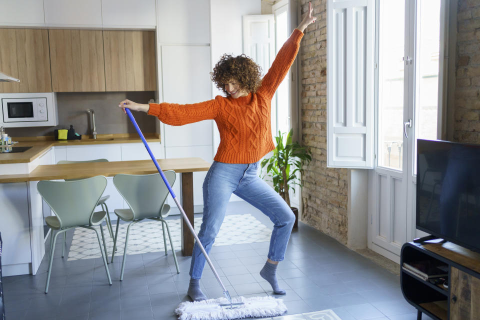 Woman in a sweater and jeans dances while mopping the floor in a modern kitchen