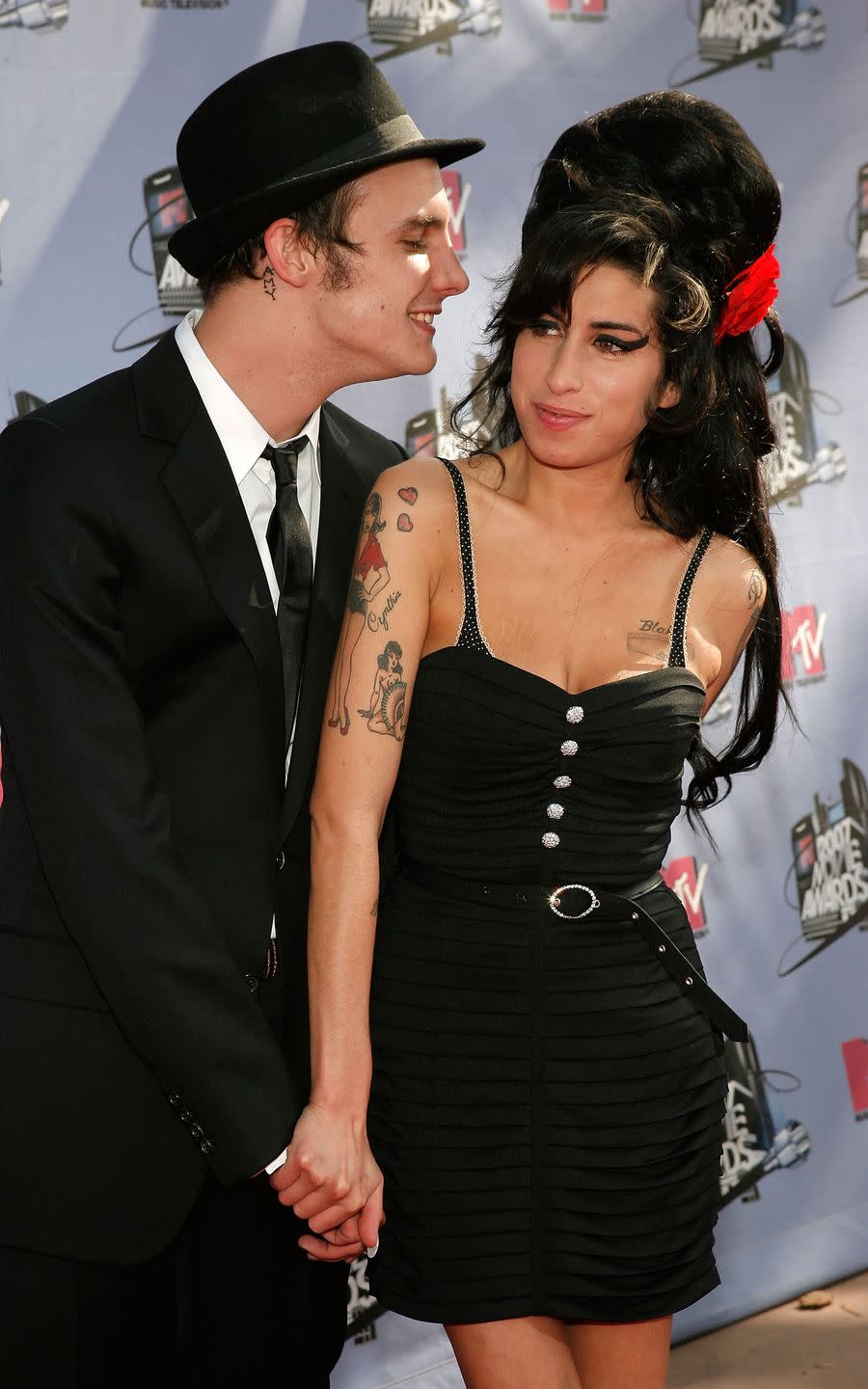 blake fielder civil and amy winehouse stand together in front of lavender photo backdrop with logos, he looks at her and smiles and she looks to the left and smiles, they hold hands and both wear black outfits