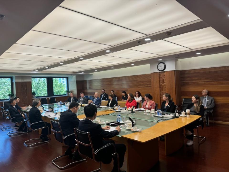 A group of Chinese government officials sit at one side of a long wooden board room table facing officials from the U.S. Dairy Council. In the background are green trees in three windows.