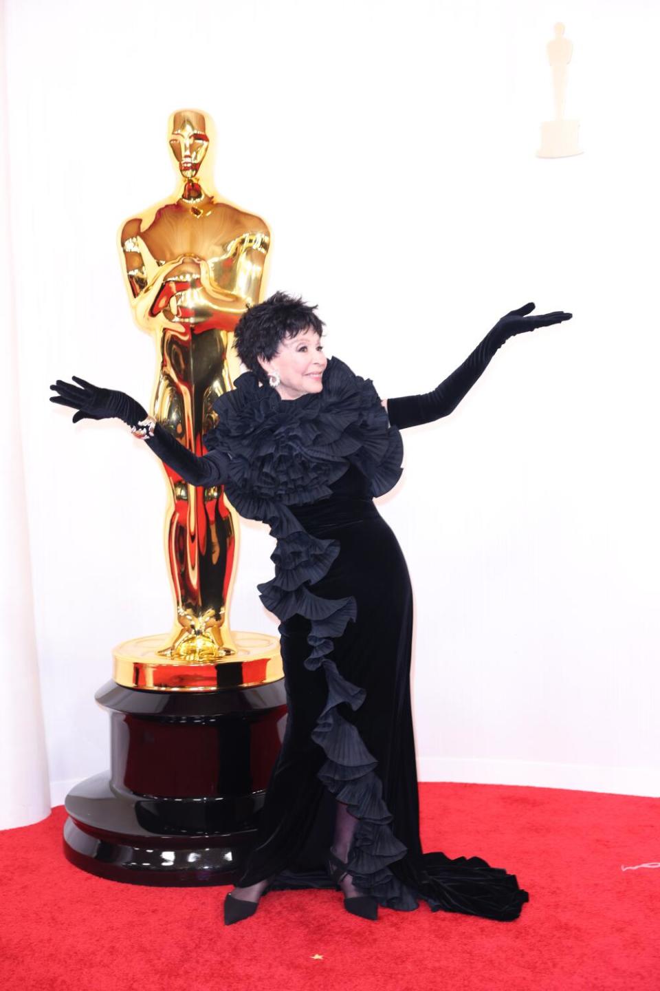 Rita Moreno, in a black dress, stands next to an Oscar statue with her arms outstretched.