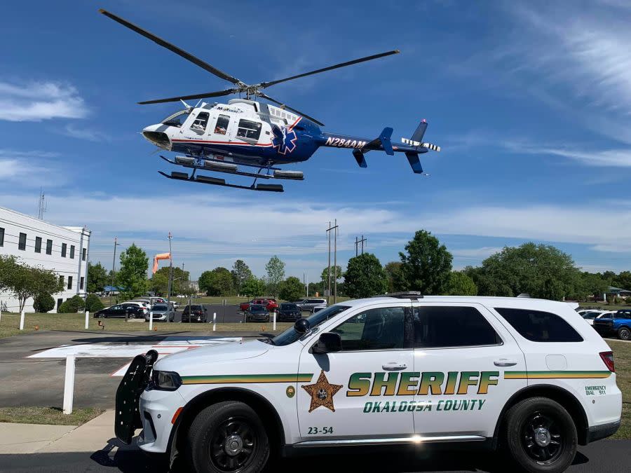 A photo of a Med Flight helicopter in the air with an Okaloosa County Sheriff's patrol car below it (OCSO).