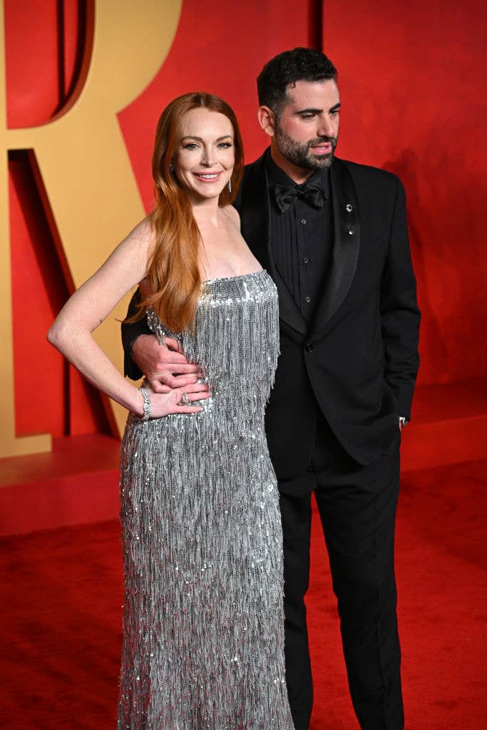 Two individuals posing on red carpet; person on left in a sleeveless, shimmering, full-length gown; person on right in classic black tuxedo