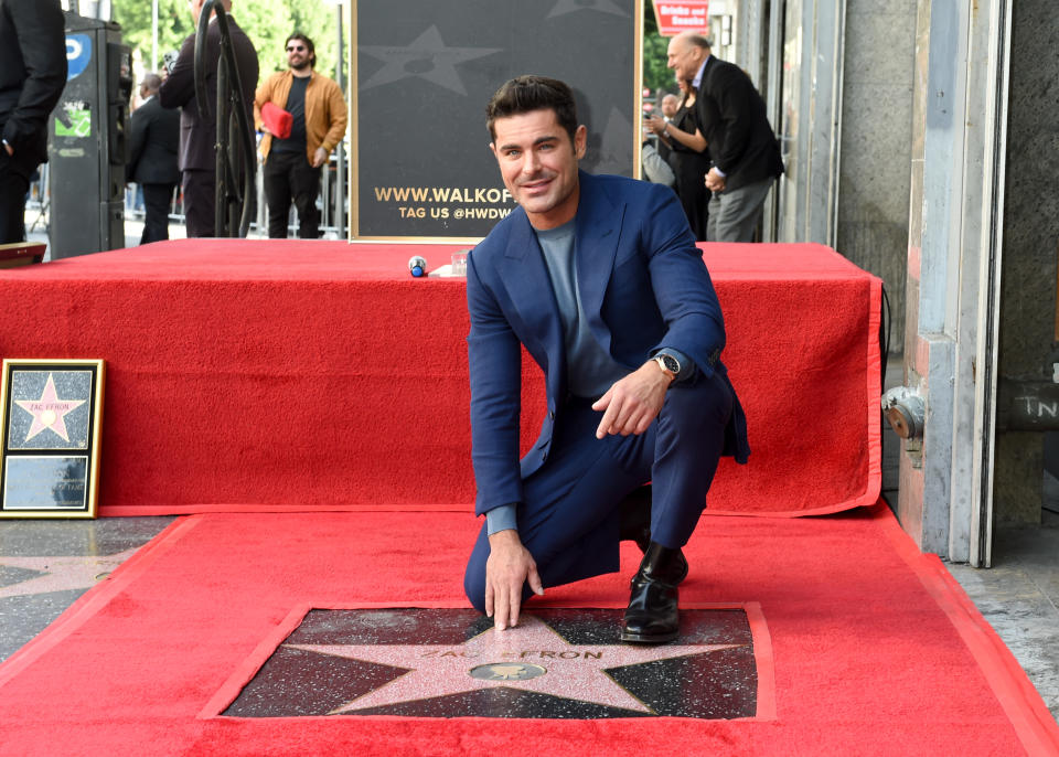 Zac Efron kneels next to his Hollywood Walk of Fame star during a ceremony. He is wearing a tailored suit and smiling at the camera