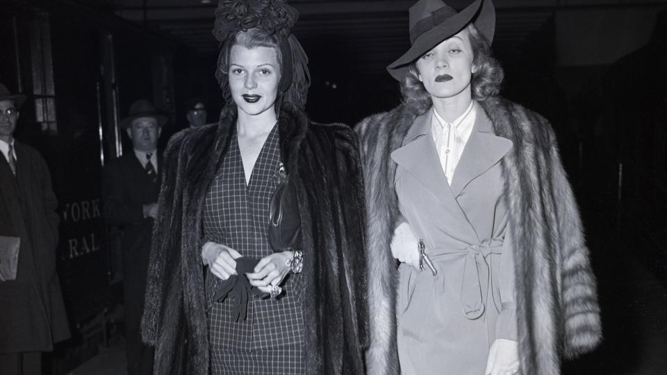 rita hayworth and marlene dietrich walk together, both wear fur jackets on their shoulders, hats, skirt suits and high heels