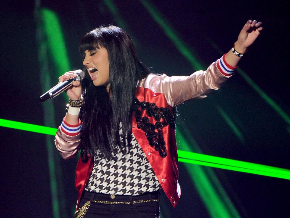 Jena Irene Asciutto wearing a white and red jacket with eyes closed singing into microphone with green lights in background