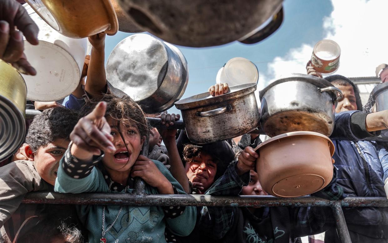 Palestinian children, holding empty pots, wait in line to receive food prepared by volunteers for Palestinian families ,displaced to Southern Gaza due to Israeli attacks, between rubbles of destroyed buildings in Rafah, Gaza