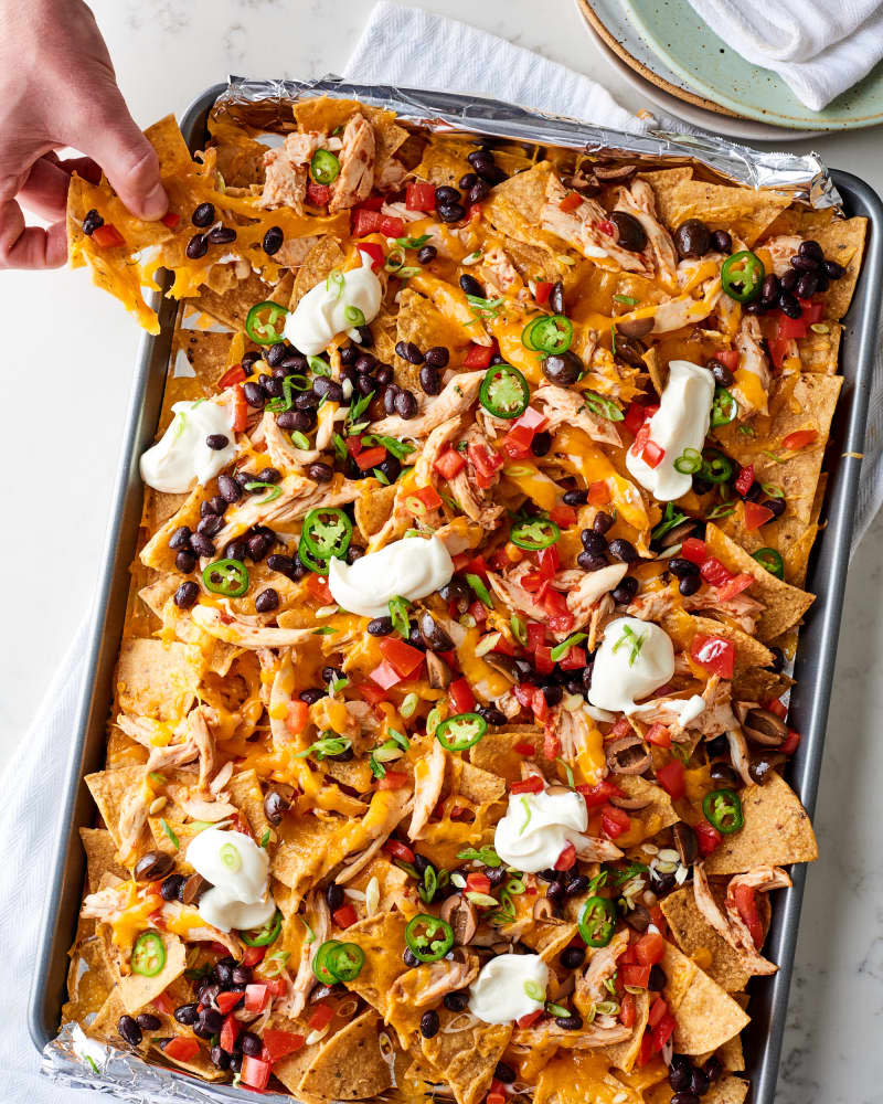 Easy sheet pan chicken nachos in the pan, one hand pulls a couple of chips out