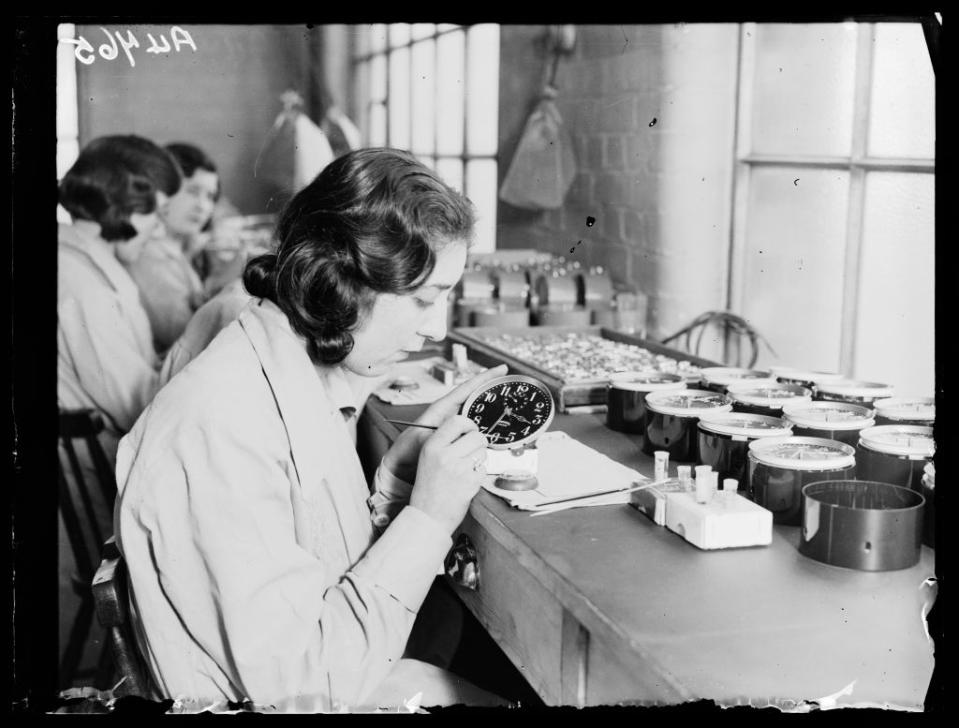 Several women are seated at a workbench assembling or inspecting clock dials in a factory setting