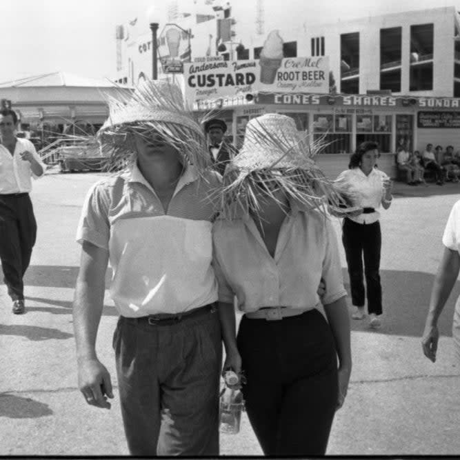 People walking at an amusement park, two wearing large straw hats covering their faces, others in casual 1950s attire