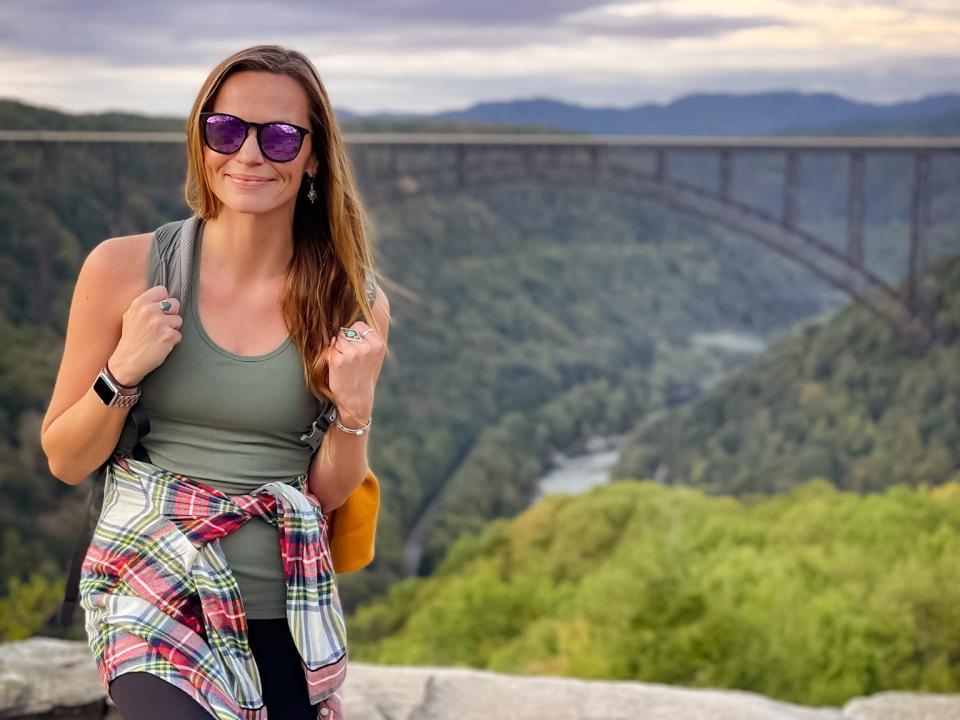 Emily, wearing sunglasses, a green tank top, black leggings, and a flannel around her waist, stands in front of New River Gorge Bridge.