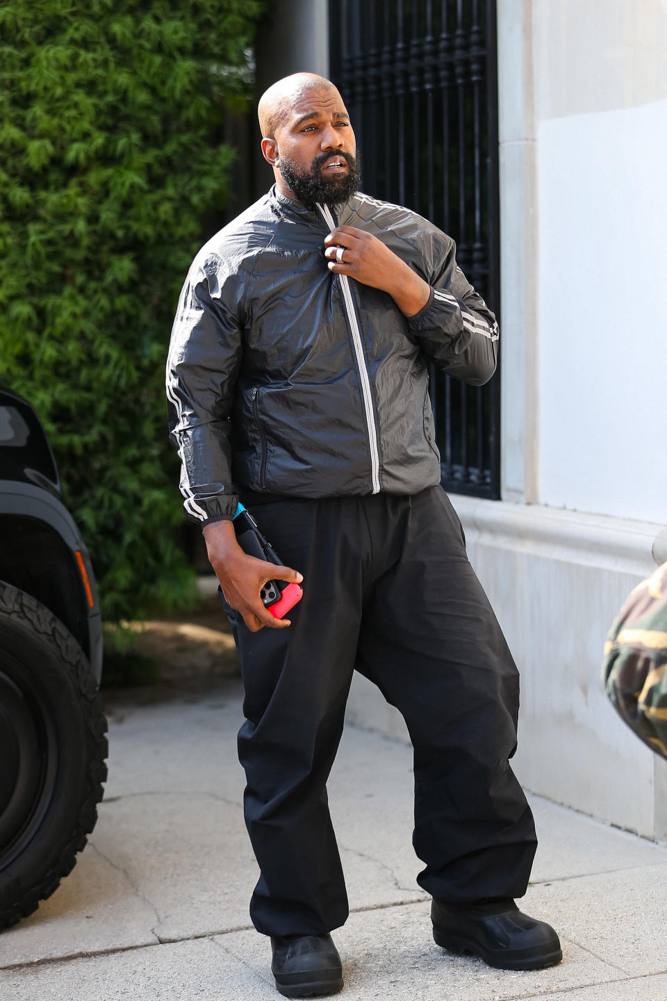 Kanye West stands outside, adjusting his black jacket. He holds a red item in his left hand and wears black pants